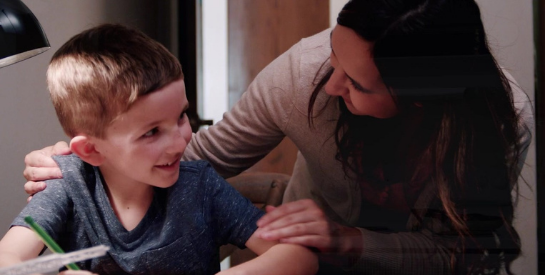 Young boy smiling at his desk with his mom looking over his shoulder with a smile