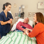 Smiling CNA standing by a older male patient's bedside, with his smiling adult daughter sitting next to his bed