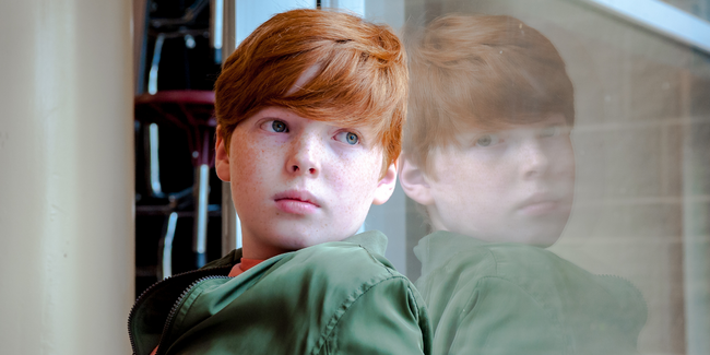 Serious looking red haired young boy looking out a window in school_Isaac